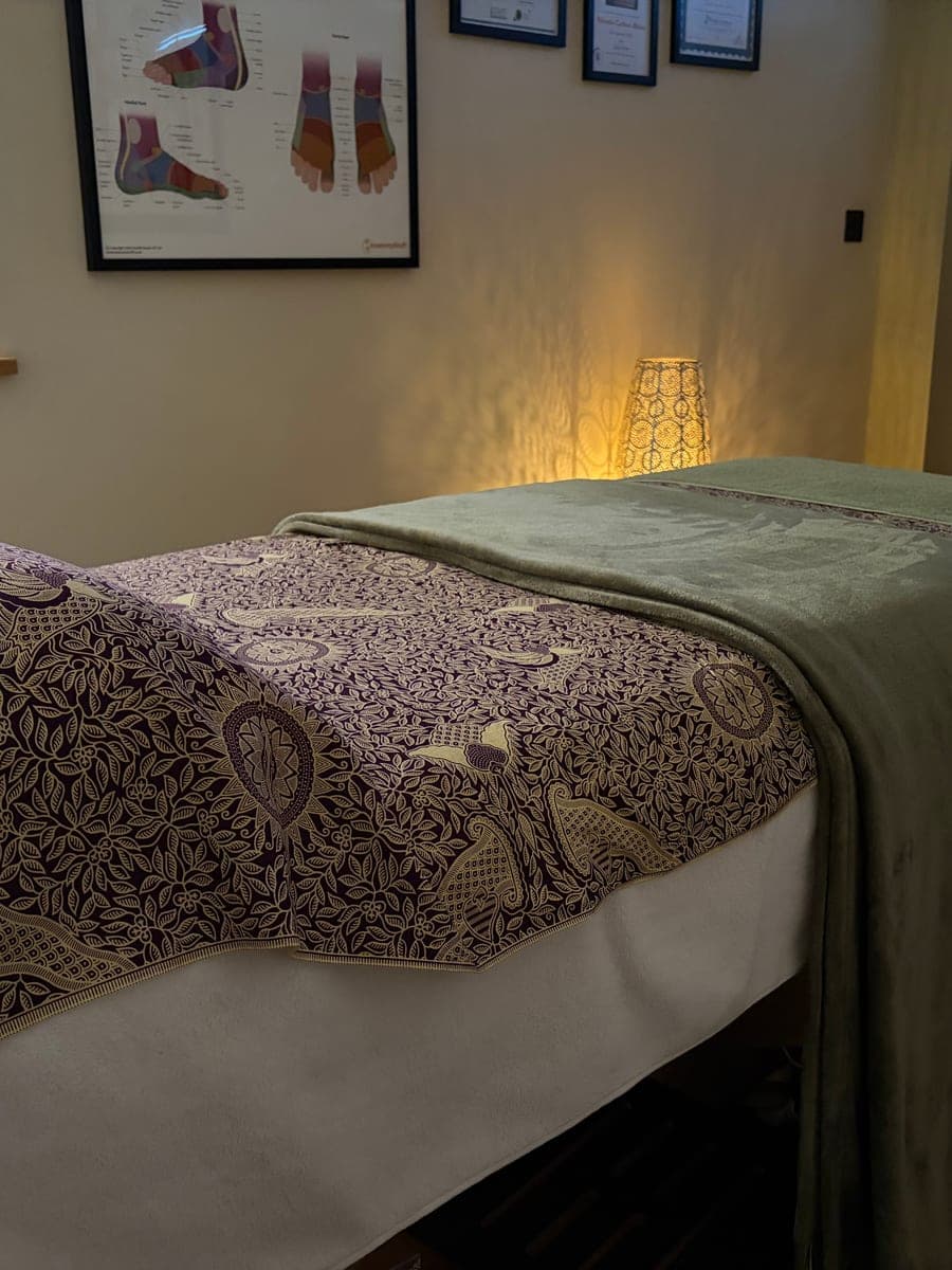 Close up of the treatment couch, made up with a patterned throw and a soft green blanket, in warm lamplight, with foot reflexology charts on the wall.