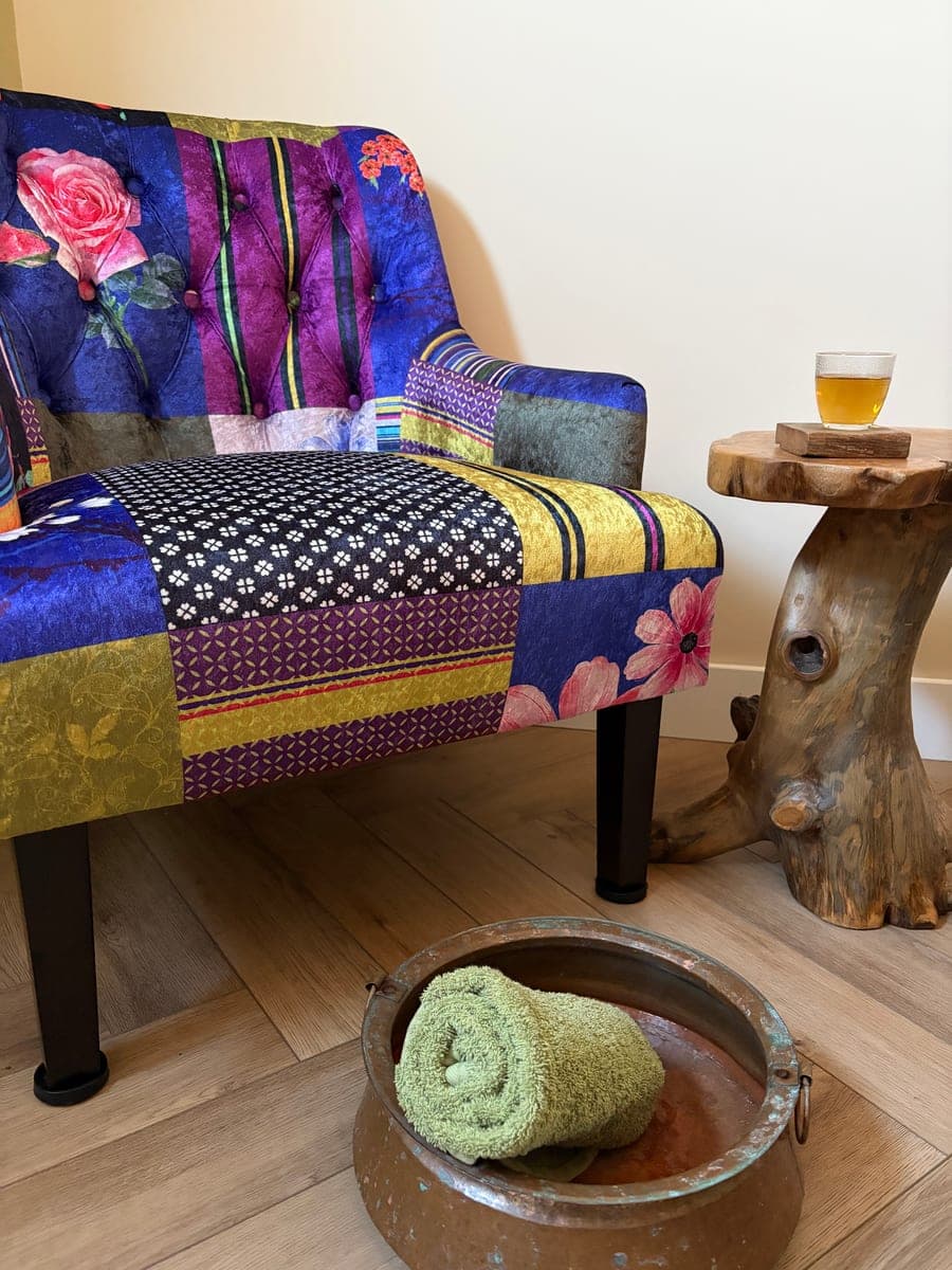 A patchwork armchair beside a small wooden table with a glass of herbal tea, and a copper foot bowl with a rolled green towel on the floor.