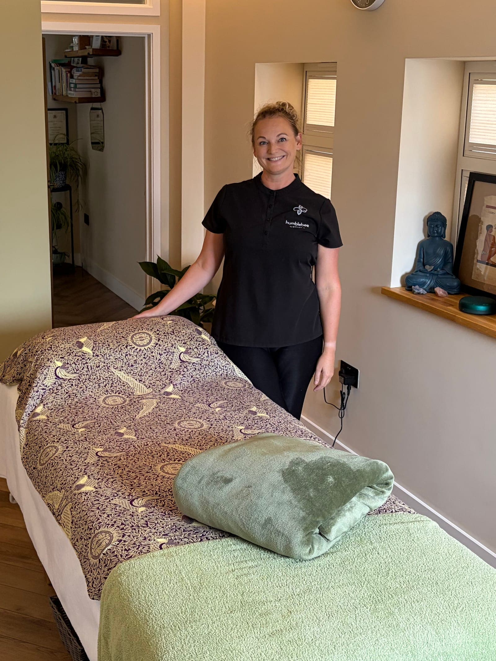 Kirstie smiling beside the made-up treatment couch in her room at Humblebee Wellness, with a Buddha figure on the windowsill.