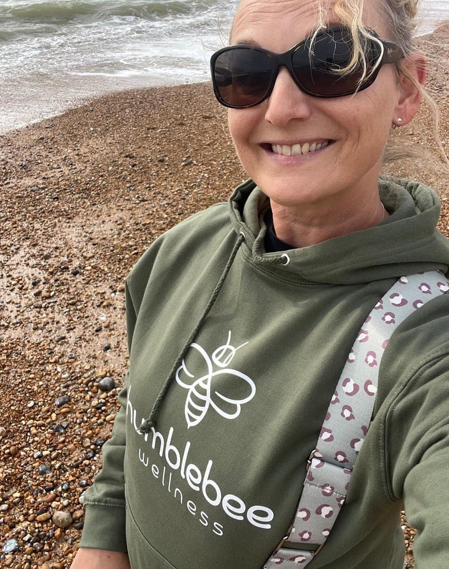 Kirstie on the shingle beach in sunglasses and a green Humblebee Wellness hoodie, the sea behind her.